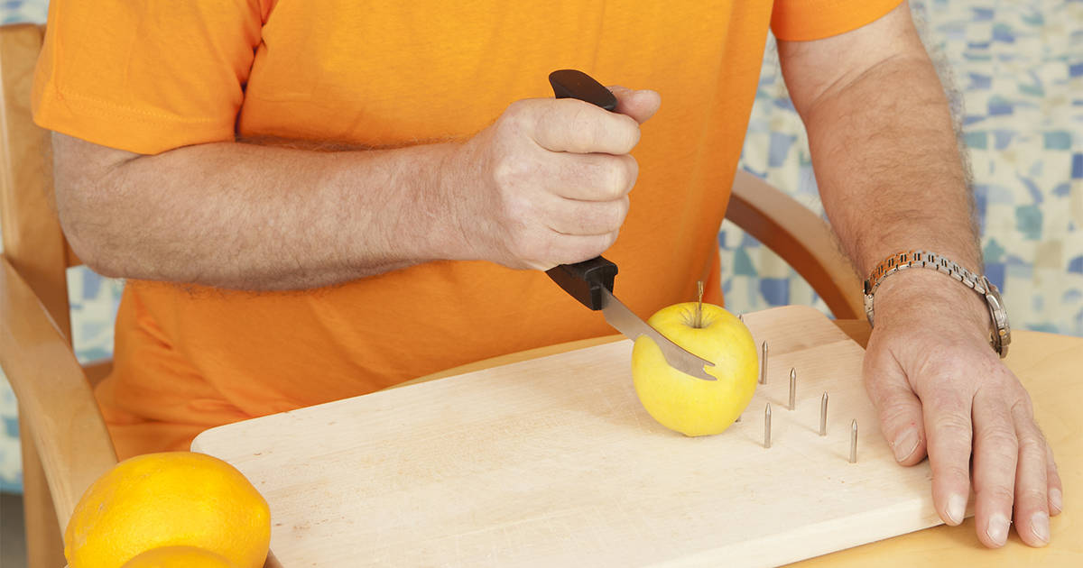 Miehen käsi leikkaa keltaista omenaa leikkuulaudalla veitsellä, jonka kahvaosa on pystyssä terään nähden
