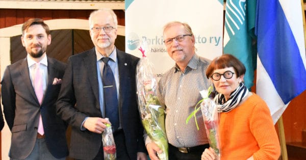 Kolme miestä ja yksi nainen seisovat rinnakkain ja hymyilevät kameralle. Kolmella on kädessään ruusut..